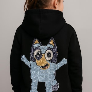 Puede incluir: Sudadera con capucha negra con un diseño de perro de dibujos animados azul claro brillante en la espalda. El perro tiene una nariz y boca amarillas, con detalles azules y morados. Ropa infantil.