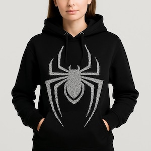Könnte beinhalten: Schwarzer Kapuzenpullover mit einem großen, glitzernden Spinnenmuster. Die Spinne besteht aus kleinen, glitzernden Elementen. Der Pullover hat eine Vordertasche und eine Kapuze mit Kordelzug. Ideal für Halloween.