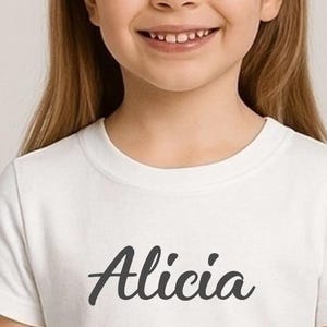 May include: A white t-shirt featuring the name "Alicia" in dark grey script. The t-shirt is worn by a young girl with long, light brown hair. The girl is smiling.