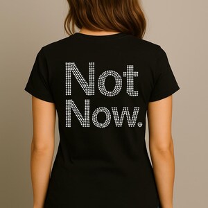 May include: Black t-shirt with the words "Not Now." in white rhinestones on the back. The shirt has a crew neck and is made of a soft fabric. The text is in a simple, clear font.