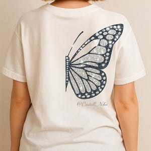 May include: White t-shirt featuring a large butterfly design on the back. The butterfly has a silver glitter pattern with a dark blue outline. The text "@Cnstall_Nika" is printed below the butterfly design.