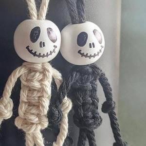 Può includere: Due figure di scheletro in macramè fatte a mano con teste di legno. Lo scheletro a sinistra è fatto con corda di colore naturale e lo scheletro a destra è fatto con corda nera. Entrambi gli scheletri hanno un viso bianco dipinto con occhi neri e una bocca cucita.