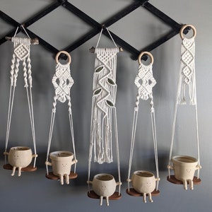 Może przedstawiać: Osiem makramowych wieszaków na rośliny z drewnianymi pierścieniami, wiszących na czarnym metalowym stojaku. Każdy wieszak ma mały ceramiczny doniczkę z twarzą i nogami, przypominającą osobę siedzącą na huśtawce.