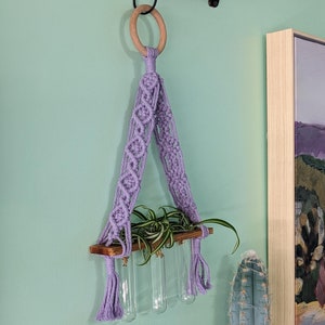 Op de afbeelding: Een handgemaakte macramé plantenhouder in lavendel, opgehangen aan een houten ring en een zwarte metalen haak. De houder heeft een houten plank met heldere glazen buizen voor planten. Een kleine groene plant is zichtbaar.