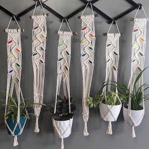 Può includere: Sette portavasi in macramè con accenti colorati appesi a un rack in metallo nero. Ogni portavasi è realizzato con cordoncino di cotone bianco e presenta un diverso motivo di foglie colorate. I portavasi sono esposti con piante in vaso.