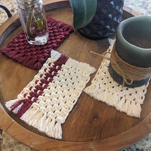 May include: A wooden tray holds several macrame coasters in burgundy and cream colors. One coaster is solid burgundy, while others have a striped design. The coasters have fringe detailing. A glass jar with a plant and a dark green candle are also on the tray.