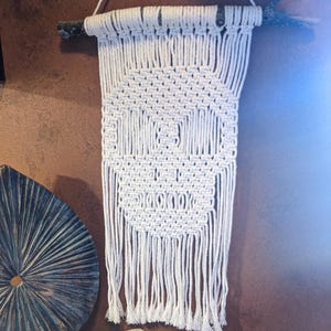 Puede incluir: Colgante de pared de macramé blanco con diseño de calavera, suspendido de una rama de madera natural. La calavera está creada con nudos cuadrados y la parte inferior tiene flecos. En primer plano, objetos decorativos con temática otoñal.