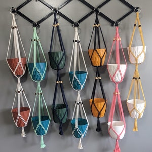 Op de afbeelding: Tien macramé plantenhangers in verschillende kleuren, waaronder wit, zwart, groen, roze en geel. Elke hanger heeft een geweven ontwerp en een kleine pot aan de onderkant. De potten zijn gemaakt van hard plastic en zijn in verschillende kleuren.