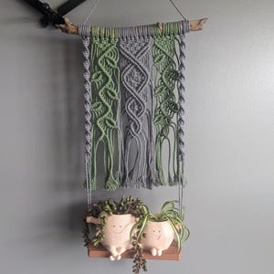 Może przedstawiać: Wiszący makramowy gobelin w kolorze szarym i zielonym z drewnianym drążkiem u góry. Gobelin jest zawieszony na suficie i ma frędzle na dole. Dwa małe ceramiczne doniczki z uśmiechniętymi buźkami wiszą na dole makramowego gobelinu.