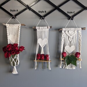 May include: Three macrame wall hangings, each with a different design. Style A holds a glass jar filled with red roses. Style B features a heart design and holds red roses on a small shelf. Style C has a heart design and holds red roses and green leaves.