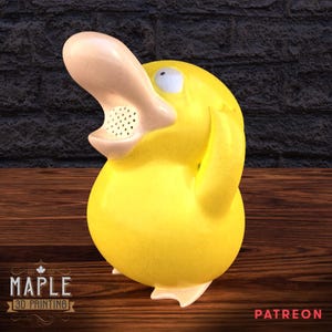 May include: A yellow 3D-printed figurine of a cartoon duck-like creature with a large, light-colored bill and a surprised expression. The figurine has a textured surface and is standing on a wooden surface. The Maple 3D Printing logo is visible.