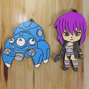 May include: Two cartoon-style character cutouts. One is a blue robot-like figure with white and gray accents. The other is a female anime character with purple hair, a black jacket, and a gun. Both have a small loop at the top for hanging.
