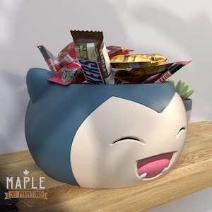 May include: A blue and white 3D-printed Snorlax-shaped container filled with various candy bars. The container has a smiling face with a pink mouth and is sitting on a wooden shelf. The Maple 3D Printing logo is visible.