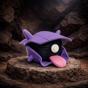 May include: A purple and black toy figure with a pink tongue sticking out. The figure has a shell-like structure on top and is set on a stone surface. The background is a rocky texture.