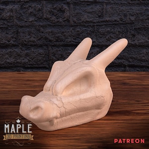 May include: A light beige dragon head sculpture with two horns and a textured surface. The dragon head is resting on a wooden surface with a dark brick background. The Maple 3D Printing logo is visible.