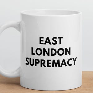 May include: A white ceramic mug with the text "EAST LONDON SUPREMACY" in black, displayed on a wooden surface. The mug has a curved handle and a cylindrical shape, suitable for hot beverages.