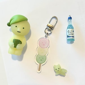 May include: A set of four green and yellow character charms. The charms are in the shape of a person holding a cup, a person with a sleeping cap, a person with a cat face, and a keychain with three round dango charms. The dango charms are pink, yellow, and green. The keychain also has a small blue bottle charm with Korean text that reads "Soju".