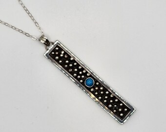 Può includere: Un ciondolo rettangolare in argento con uno sfondo scuro e testurizzato riempito di piccole sfere argentate e una singola pietra rotonda blu brillante al centro. Il ciondolo è appeso a una sottile catena a maglie d'argento.