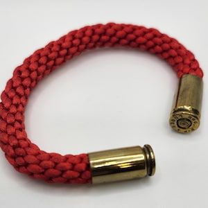 May include: Red braided bracelet with brass bullet casing closures. The bracelet is made of a thick, woven red cord. The bullet casings are a shiny gold color and are engraved with text.