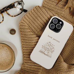 Könnte beinhalten: Cremefarbenes Handycase mit dem Schriftzug "book person" und einer floralen Illustration eines offenen Buches. Das Case liegt auf einem braunen Strickpullover, im Hintergrund eine Brille und eine Kaffeetasse.