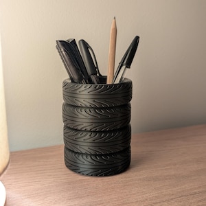 May include: Black tyre-shaped pencil holder with five pens and pencils inside. The holder is made of black plastic and has a tyre tread pattern.
