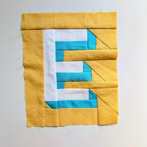 May include: A square fabric block with a patchwork design. The block has a yellow border, white and turquoise stripes, and geometric shapes. The fabric has a textured appearance, possibly for a quilt or craft project.