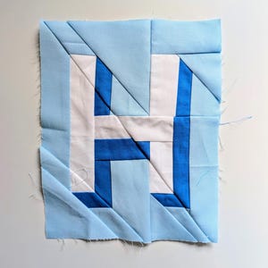 以下が含まれることがあります： 水色の背景に、白と青の「H」の文字がデザインされた正方形の布ブロック。文字は幾何学的な形で構成されており、青色の要素が文字の形を形成しています。ブロックの縁も水色です。