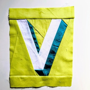 May include: A square fabric piece with a lime green border and a central V-shaped design. The V is composed of white and teal fabric sections, creating a geometric pattern. The piece is likely part of a larger textile project.