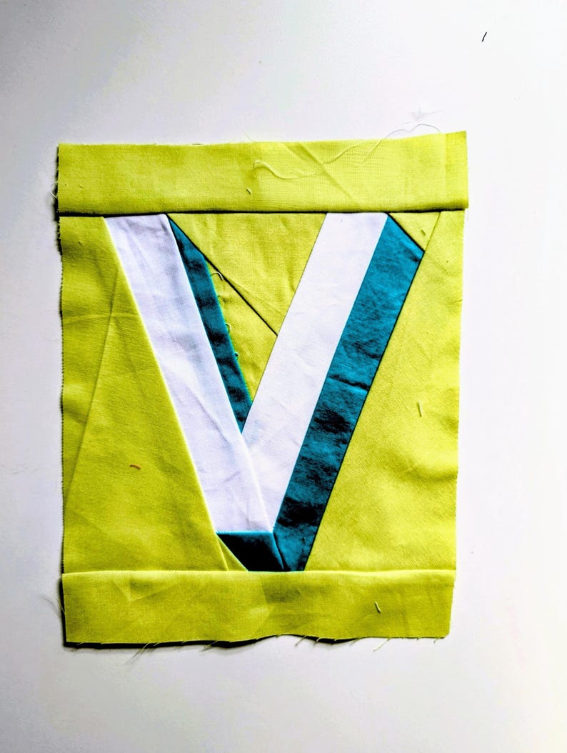 May include: A square fabric piece with a lime green border and a central V-shaped design. The V is composed of white and teal fabric sections, creating a geometric pattern. The piece is likely part of a larger textile project.