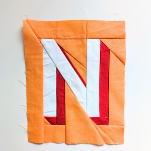 Pode incluir: Um bloco de tecido quadrado com uma borda laranja e um design de letra "N" branco e vermelho. A letra é composta por pedaços de tecido brancos e vermelhos, criando um padrão geométrico. O bloco provavelmente faz parte de um projeto têxtil maior.