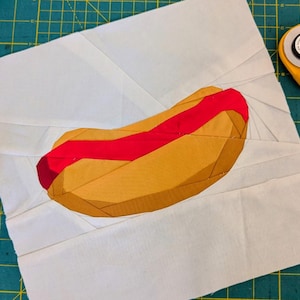 May include: A fabric art piece featuring a stylized hot dog. The hot dog has a red sausage and a yellow bun, set against a white background. The piece is on a green cutting mat, with a rotary cutter and ruler visible.