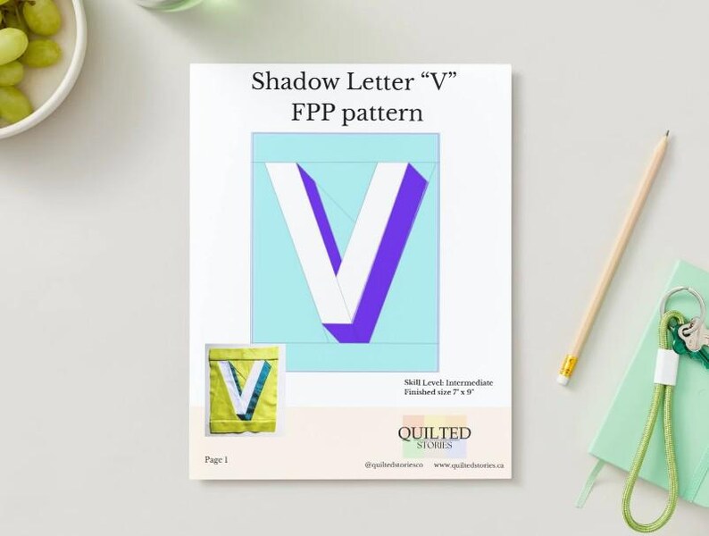 May include: A white paper with the text "Shadow Letter V FPP pattern" and a large letter "V" in white and purple. A smaller image of the letter "V" is in the lower left corner. A pencil and a green notebook are on the right.
