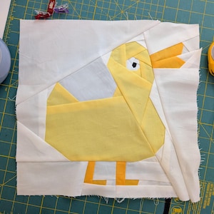 May include: A patchwork quilt block depicting a yellow duck. The duck is crafted from yellow fabric with orange feet and beak, set against a white and light grey backdrop. The block rests on a green cutting mat.