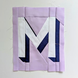 May include: A handmade fabric block featuring the letter "M" in a patchwork design. The letter is composed of white, navy blue, and lavender fabric pieces. The block has a lavender border and is likely part of a larger quilt or craft project.