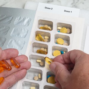 May include: A person's hand holding several orange softgel capsules next to a pill organizer filled with various pills. The organizer is labeled with "Dose 1" and "Dose 2" and the days of the week.