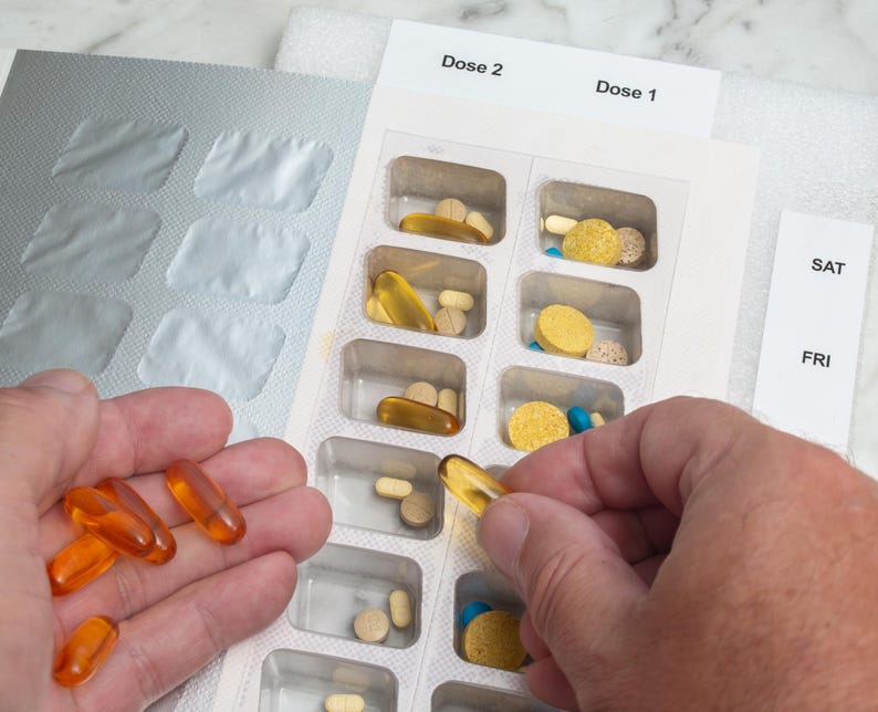 May include: A person's hand holding several orange softgel capsules next to a pill organizer filled with various pills. The organizer is labeled with "Dose 1" and "Dose 2" and the days of the week.