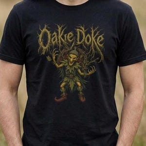 Pode incluir: T-shirt preta com o texto dourado "Oakie Doke" e uma ilustração detalhada de uma figura esquelética com roupa de folhas e sapatos vermelhos. A figura tem os braços estendidos. A t-shirt é feita de um material macio.