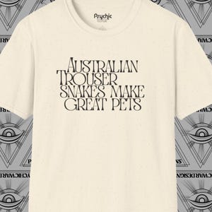 May include: A cream-coloured t-shirt featuring the text "AUSTRALIAN TROUSER SNAKES MAKE GREAT PETS" in black. The shirt has a crew neck and short sleeves. The brand name "Psychic War" is printed at the top.