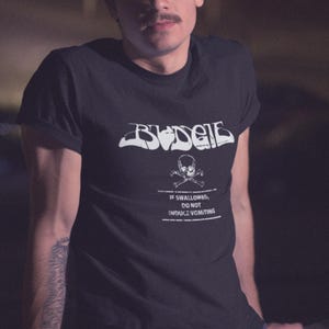 May include: Black t-shirt with a white graphic of the word "Budgie" in a stylized font. Below the word is a skull and crossbones with the text "If swallowed, do not induce vomiting."