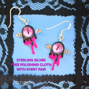 Puede incluir: Un par de pendientes colgantes de plata de ley. Cada pendiente presenta un diseño negro y rosa con un detalle central en forma de ojo y pequeñas alas plateadas. Los pendientes tienen un estilo artístico único.