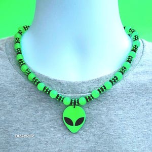 Op de afbeelding: Een ketting met een fel neongroene alienhoofd-hanger met zwarte ogen, geregen aan een koord met afwisselend neongroene ronde kralen en kleine zwarte tussenkralen. Het woord DIZZYPOP staat in het wit gedrukt in de linkerbenedenhoek.