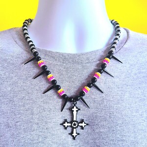 Puede incluir: Un collar de cuentas blanco y negro con un colgante de cruz. El collar presenta púas negras y cuentas de colores en rosa, amarillo y negro. El colgante de cruz tiene una estrella en el centro.