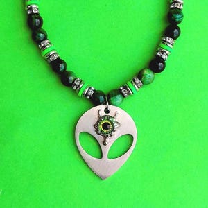 Op de afbeelding: Een unieke ketting met een alien hanger. De hanger is zilverkleurig met een oogvormig midden. De ketting heeft groene en zwarte kralen, met zilverkleurige afstandhouders. De achtergrond is felgroen.