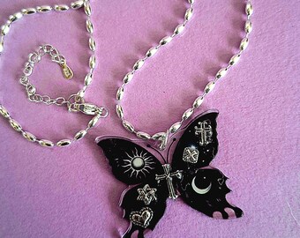 Puede incluir: Collar plateado con un colgante de mariposa negro. El colgante de mariposa presenta símbolos celestiales y religiosos, incluyendo un sol, una luna, cruces y estrellas. El collar está sobre una superficie púrpura.