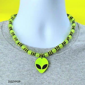 Op de afbeelding: Een ketting met een limoengroene alien-hoofdhanger met zwarte ogen. De ketting bestaat uit doorschijnende limoengroene ronde kralen afgewisseld met kleinere zwart-geel gepatroonneerde cilindrische kralen. De tekst DIZZYPOP is zichtbaar.