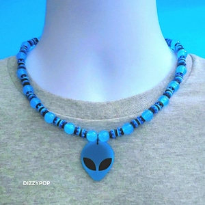 Puede incluir: Un collar con un colgante de cabeza de alienígena azul con ojos negros, ensartado en un cordón con cuentas redondas de color azul translúcido y pequeños espaciadores negros. El texto DIZZYPOP está impreso en blanco en la parte inferior.
