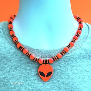 Könnte beinhalten: Eine Halskette mit einem leuchtend orangefarbenen Alien-Kopf-Anhänger mit schwarzen Augen. Die Kette besteht aus abwechselnden matten orangefarbenen Kugeln und kleineren schwarzen, strukturierten Zwischenperlen. Der Text DIZZYPOP steht unten links in Weiß.