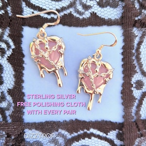 May include: A pair of gold-colored heart-shaped earrings with a pink center and a dripping design. The earrings have a chain design around the heart. The text "STERLING SILVER FREE POLISHING CLOTH WITH EVERY PAIR" is visible.