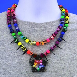 Puede incluir: Un collar de cuentas de colores con un arcoíris de colores, púas negras y un colgante en forma de flor. El collar se muestra en un maniquí.