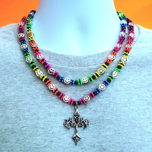 Puede incluir: Un collar de cuentas de doble capa con cuentas plateadas de cara sonriente intercaladas con cuentas de color neón rosa, naranja, amarillo, verde y azul, con un colgante central de cruz plateada ornamentada en la hebra inferior.